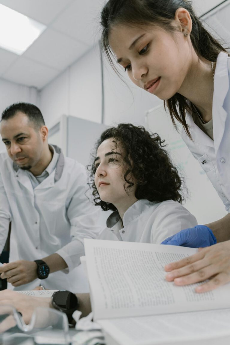 Three scientists wearing lab coats collaborate on research with books and computers in a modern laboratory setting.